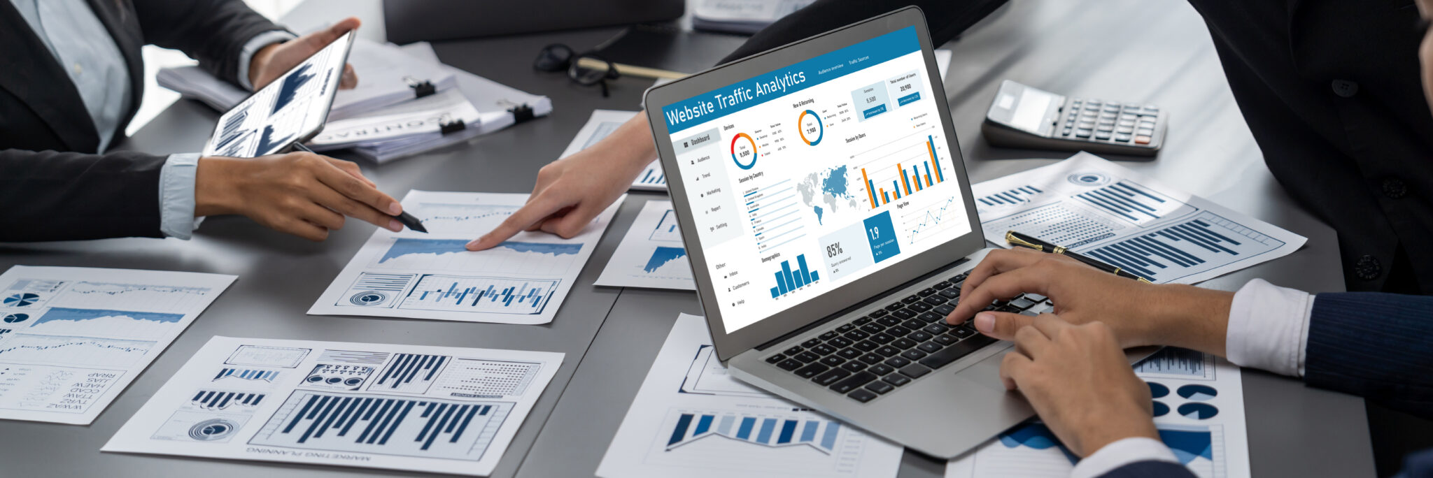 A website traffic analytics dashboard open on a laptop while two people review printed charts on the desk
